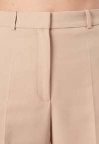 Beige tailored trousers with a smooth texture, featuring a concealed front pocket and subtle stitching details along the waistline.