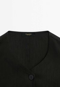 Svart blus med v-ringning, jämn textur, subtil lyster och enstaka knäppning. Varsel om varumärke syns vid halsringningen.