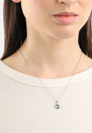 Collier en argent avec un pendentif en forme de goutte d'eau bleu, porté sur une chemise rayée beige clair, avec une chaîne délicate et une texture lisse.