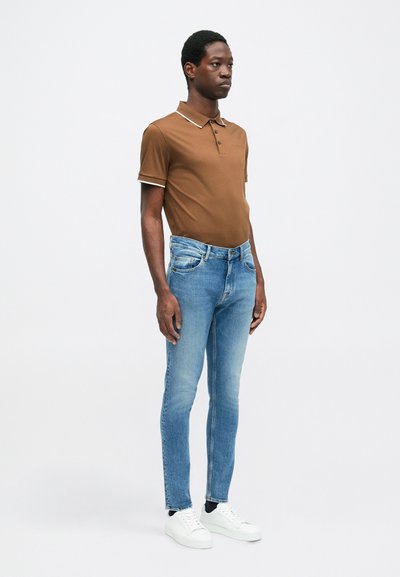 Camisa polo marrón con detalles blancos, jeans ajustados azules y zapatillas blancas. El modelo está de pie contra un fondo claro y liso.