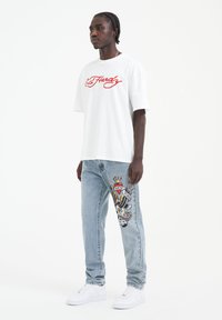 T-shirt bianco oversize con logo rosso. Jeans azzurri chiari con grafica colorata sulla gamba sinistra. Sneakers bianche completano l'outfit.