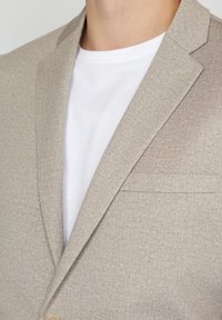 Beige blazer in textured fabric with notched lapels, single chest pocket, and a button closure, worn over a white t-shirt.