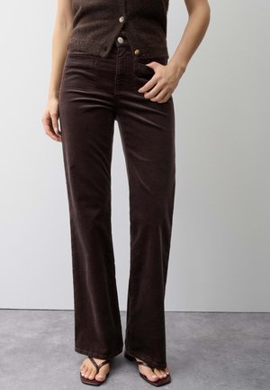 Femme portant un pantalon évasé taille haute marron foncé avec poches avant, un haut sans manches boutonné et des sandales marron, debout sur un sol gris.