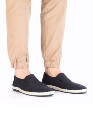 Schwarze Slipper mit perforierter Textur, weißer genähter Sohle und elastischen Seitenpaneelen. Getragen mit hellbraunen Khakihosen.