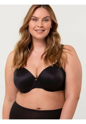 Uśmiechnięta kobieta z długimi falującymi włosami, ubrana w czarny biustonosz z fiszbinami i pasujące czarne majtki na jednolitym tle.