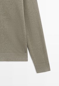 Section d'un pull en tricot vert olive à manches longues montrant les poignets et l'ourlet côtelés sur un fond blanc.