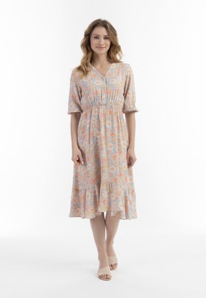 Blumenkleid in Pastellfarben mit kurzen Puffärmeln, geraffter Taille und Knopfdetails. Getragen mit hellen offenen Schuhen.