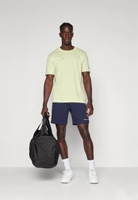 Gaiši zaļa īsroku T-krekls ar "Champion" logotipu, tumši zili šorti, balti sporta apavi, tur melnu sporta soma. Gluda auduma, ikdienišķa piegriezuma.