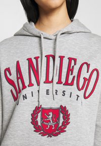 Felpa grigia con testo rosso "Università di San Diego" e uno stemma rosso che presenta un leone e una corona di alloro, indossata da una persona con capelli neri lisci.