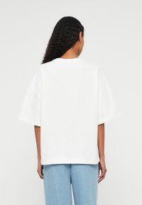 Λευκό υπερμέγεθες βαμβακερό T-shirt με κοντά μανίκια και στρογγυλή λαιμόκοψη, βλέπεται από πίσω, συνδυασμένο με ανοιχτό γαλάζιο τζιν παντελόνι.