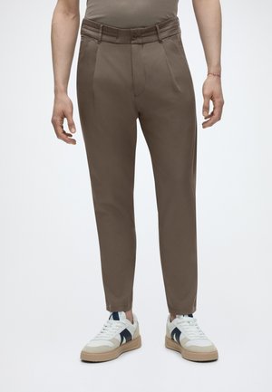 Mann trägt braune Slim-Fit-Hose und weiße Sneakers mit schwarzen Details, steht vor einem schlichten hellen Hintergrund.