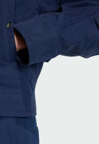 Námořnická modrá bunda s texturovanou tkaninou, zapínáním na knoflíky a detailním prošíváním na rukávech. Design zdůrazňuje silný a pevný materiál.