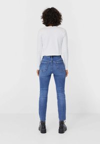 Haut blanc léger à manches longues associé à un jean bleu taille haute, coupe slim, avec poches plaquées et texture lisse. Des bottes noires complètent la tenue.
