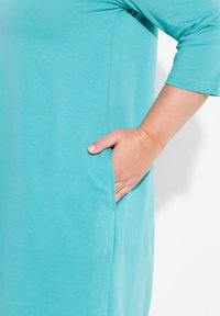 Een turquoise jurk met driekwartmouwen, voorzien van een zijzak. De stof lijkt zacht en glad, met een relaxte pasvorm.