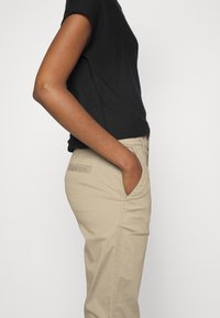Personne en pantalon beige avec la main dans la poche, portant un haut noir à manches courtes sur un fond blanc uni.