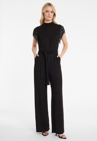 Zwarte jumpsuit met korte mouwen, voorzien van hangende zwarte franjes, een hoge halslijn, een strikceintuur in de taille en wijde broekspijpen.