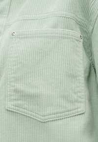 Tissu en velours côtelé vert clair avec une poche carrée cousue ornée de deux boutons-pression argentés aux coins supérieurs.