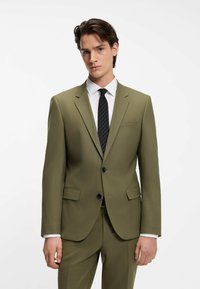 Traje verde olivo de corte ajustado, cierre de un solo botón, solapas con muesca y botones negros. Combinado con camisa blanca y corbata negra a rayas.