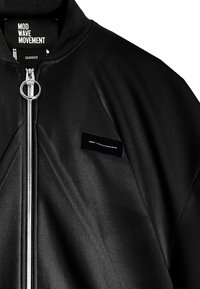 Chaqueta bomber negra con cremallera frontal y tirador redondo; presenta una pequeña etiqueta negra, tejido texturizado y un sutil detalle de acolchado.