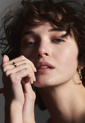 Les bijoux en or comprennent une bague texturée et des boucles d'oreilles pendantes avec des accents colorés. Le sujet a les cheveux courts et ébouriffés, ainsi qu'un maquillage neutre.