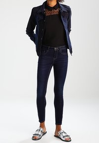 Veste en jean à boutons sur le devant, bleu foncé. Haut noir avec encolure à motifs. Jean slim foncé et sandales plates ornées d'argent.