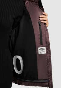 Person iført sort sweater holder åbent en brun jakke, der viser sort inderfor med lynlåslomme og et mærke, der læser "En varm kram lavet af stof."