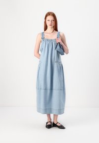 Denim maxi šaty vo svetlom modrom odtieni s nariaseným výstrihom, nastaviteľnými ramienkami a vreckami; siahajú pod kolená, bezšvíkový lem, v kombinácii s čiernymi topánkami.
