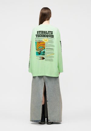 Osoba o długich brązowych włosach, ubrana w miętowo-zieloną bluzkę z długim rękawem z kolorowymi grafikami surfera oraz wyblakłą niebieską długą spódnicę dżinsową z rozcięciem z tyłu.