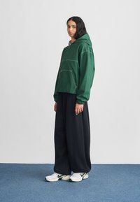 Sweat-shirt à capuche vert avec des coutures blanches contrastantes, associé à un pantalon noir ample et des baskets blanches avec des accents verts.