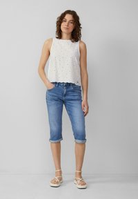 Haut sans manches blanc avec un motif à œillets et un ourlet ondulé, associé à un jean capri en denim bleu clair retroussé et des sandales beiges à lanières.