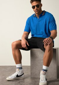 Modrá polo tričko s límečkem ve oranžové a černé barvě, kombinované s černými šortkami. Bílé tenisky s černými akcenty a pruhovanými ponožkami.