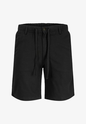 Teenager-Junge mit lockigem Haar, der einen beigen Sweatshirt, schwarze Shorts, weiße Socken und weiße Sneakers trägt und vor einem weißen, schlichten Hintergrund steht.
