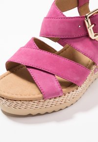 Sandal con zeppa in suede rosa, con cinturini incrociati, una soletta imbottita e dettagli in corda intrecciata simili al giute sulla suola. Presenta un accento con fibbia dorata.