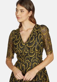 Schwarzes Kleid mit aufwendigen goldenen Blumen-Spitzenmustern, V-Ausschnitt und kurzen Spitzenärmeln mit gewellten Kanten.