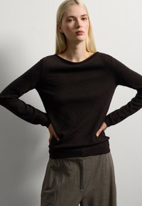 Maglione nero a maniche lunghe con scollatura rotonda e polsini a costine, realizzato in un tessuto morbido e strutturato; indossato con pantaloni grigi chiari.