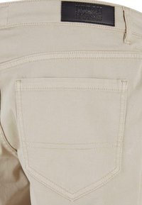 Pantalones de mezclilla beige con un solo bolsillo trasero, con una etiqueta de cuero oscura cosida en la cintura y líneas de costura visibles.