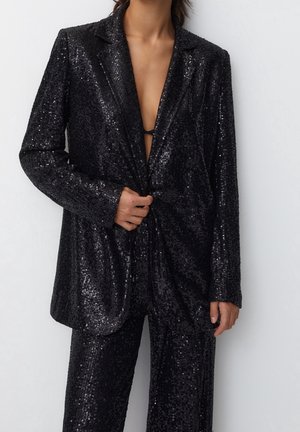 Vrouw draagt een zwarte blazer met pailletten en bijpassende broek, met een diepe halslijn en hand die het voorpand van het jasje vasthoudt.