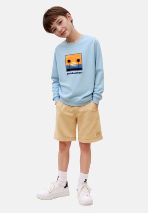 Light blue sweatshirt with palm tree graphic and "Jack & Jones" text, paired with beige shorts and white sneakers with black accents.