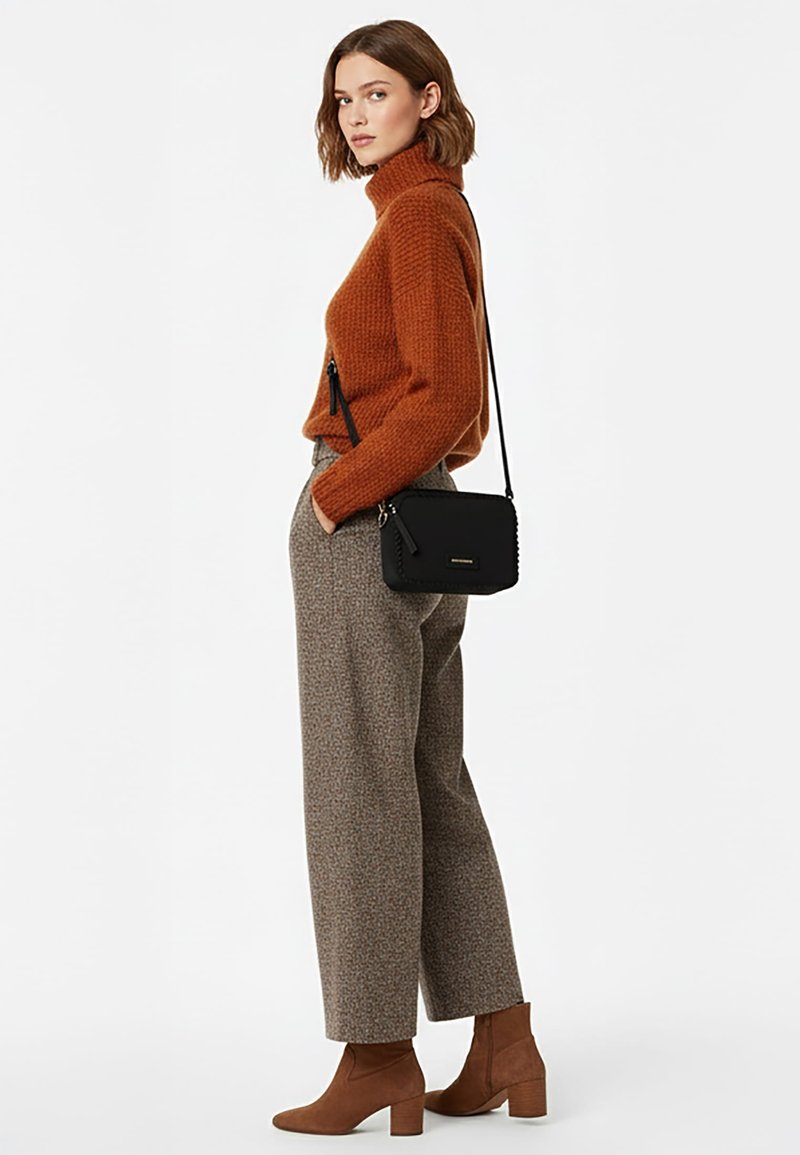 Jeune femme aux cheveux mi-longs portant un pull à col roulé couleur rouille, un pantalon gris à jambes larges, des bottines marron et un sac bandoulière noir.