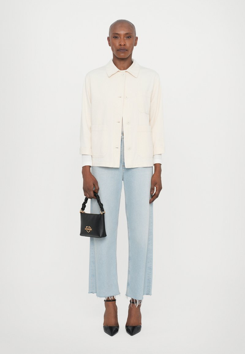 Giacca button-up crema chiara, jeans a gamba larga e strappati blu chiaro, borsa nera con manico ritorto e décolleté neri a punta.