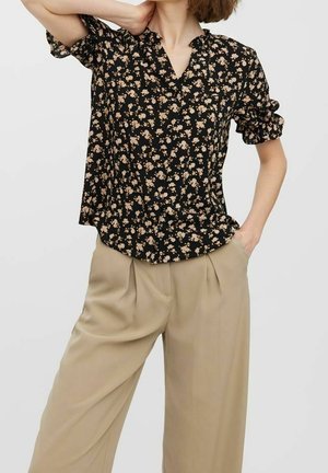 Nainen pukeutuneena mustaan, kukalliseen lyhythihaiseen V-kaulaiseen paitaan ja beigenvärisiin leveälahkeisiin laskostettuihin housuihin, poseeraa käsi kaulalla ja toinen taskussa.