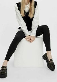 Zwarte vest over een witte shirt, gecombineerd met zwarte leggings en stevige zwarte loafers met gouden hardware. Zittend op een witte kubus.