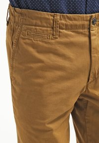 Gros plan sur une personne portant un pantalon couleur moutarde avec un bouton et des passants pour ceinture, assorti à une chemise bleu marine ornée de petits motifs blancs.