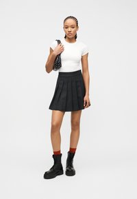 Νεαρή γυναίκα με πλεγμένα μαλλιά φοράει λευκό στενό μπλουζάκι, μαύρη πλισέ φούστα, μαύρα στρατιωτικά μποτάκια, κόκκινες κάλτσες και κρατά μια μαύρη τσάντα με τρουκς.