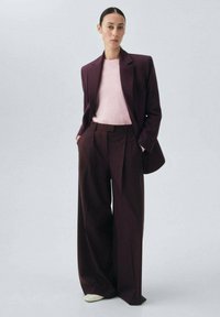 Veste de costume ajustée burgundy sur un pull rose clair, accompagnée de pantalons larges. Texture lisse, design structuré et chaussures neutres.