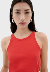 Top sin mangas de rayas rojas con un escote alto y tirantes delgados; se lleva acompañado de pendientes circulares brillantes. Textura de tela suave.