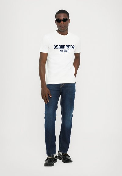 Valge T-särk, millel on tumesinine "DSQUARED2 MILANO" logo, paaritud tumedate siniste teksade ja mustade jalanikega, modell kannab prille.