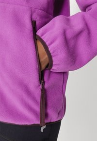 Purple fleece jacket with brown trim, featuring a zippered pocket and elastic cuffs. The material appears soft and textured.