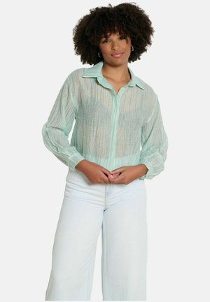 Žena s kučeravými vlasmi v svetlozelenom priesvitnom blúzke a bielych nohaviciach s vysokým pásom, stojaca s voľne zloženými rukami.