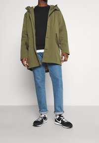 Parka oliva con cappuccio, felpa nera sotto, jeans blu e sneakers nere con accenti bianchi. Design casual e moderno.